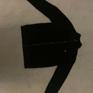 Svart fleecejacka från Lyle & Scott - Säljer en svart fleecejacka från Lyle & Scott Junior. Jackan har en dragkedja framtill och ett litet broderat emblem på bröstet. Den har ett lurvigt material så är då perfekt för kyliga dagar. 