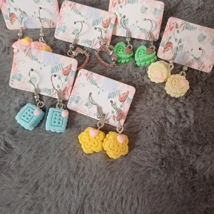 Färgglada örhängen i olika former - Säljer ett set med färgglada örhängen i olika former och färger. Setet inkluderar örhängen formade som hjärtan, kakor och blommor i färgerna blå, gul, grön och rosa. Perfekt för att lägga till en lekfull touch till din outfit!