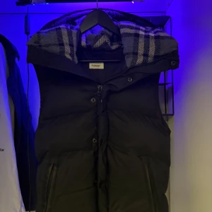 Svart dunjacka från Burberry - Snygg svart dunjacka från Burberry med en klassisk rutig insida på huvan. Jackan har dragkedja och knappar framtill samt två fickor med dragkedja. Perfekt för kyliga dagar.