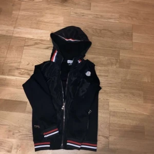 Mocler windbreaker - Snygg svart jacka från Moncler med huva och dragkedja. Jackan har ribbade muddar och en broderad logotyp på ärmen. Den har även en praktisk ficka med dragkedja på ärmen.