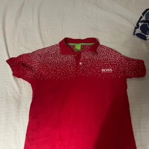 Röd kortärmad pikétröja från Boss - Snygg röd pikétröja från Boss med ett unikt vitt prickmönster över axlarna. Tröjan har en klassisk krage och korta ärmar, perfekt för en stilren look. Den gröna etiketten ger en cool kontrast. Perfekt för dig som vill sticka ut!