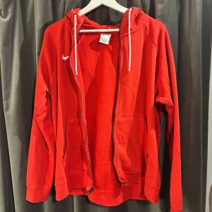 Röd Nike hoodie med dragkedja  - Säljer en snygg röd Nike hoodie med dragkedja och vit logga på bröstet. Tröjan har en justerbar huva med vita snören och praktiska fickor framtill. Perfekt för en sportig look eller avslappnade dagar. Har en lös söm på höger arm. Skriv gärna om eventuella frågor.🌺❤️