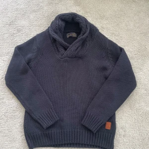 Mörkblå stickad tröja från Scotch & Soda - Säljer en snygg mörkblå stickad tröja från Scotch & Soda med en mysig krage. Tröjan har långa ärmar och ribbstickade muddar vid ärmslut och nederkant. Perfekt för kyliga dagar!