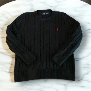 Kabelstickad Polo Ralph Luaren tröja - Säljer en stilren svart stickad tröja från Ralph Lauren med det klassiska röda logotypen på bröstet. Tröjan har en ribbad struktur och långa ärmar, perfekt för kyligare dagar. Passar både till vardags och mer uppklädda tillfällen. Stor i storleken. Passar M!