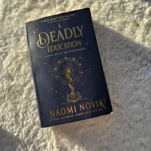 A Deadly Education - Säljer boken 'A Deadly Education' av Naomi Novik. Den har en mörkblå omslag med gyllene text och illustrationer. Perfekt för fantasyälskare som gillar magiska äventyr och spänning. En Sunday Times bestseller som fångar läsarens intresse från första sidan.