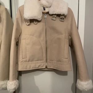 Beige jacka med fuskpäls från H&M - Säljer en snygg beige jacka från H&M med mjuk fuskpäls på kragen och ärmsluten. Jackan har dragkedja framtill och dekorativa spännen vid kragen för extra stil. Perfekt för kyliga dagar!