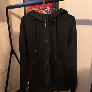 Svart hoodie med dragkedja från C.P. Company - Säljer en svart hoodie från C.P. Company med dragkedja och huva. Den har coola detaljer som glasögon på huvan och praktiska fickor med dragkedja. Perfekt för en stilren och avslappnad look.