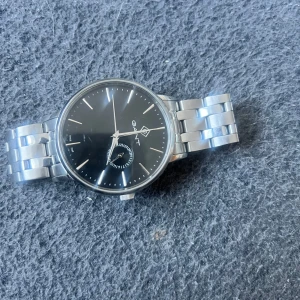 Elegant klocka från GANT - Snygg klocka från GANT med svart urtavla och silverfärgad metallarmband. Klockan har minimalistisk design med tydliga visare och en liten sekundvisare. Perfekt för den stilmedvetna som vill ha en tidlös accessoar.