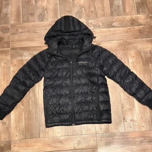 Svart pufferjacka från Peak Performance - Säljer en svart pufferjacka från Peak Performance. Jackan har en dragkedja framtill och en justerbar huva. Perfekt för kyliga dagar med sin quiltade design som håller värmen. Den har långa ärmar och en stilren look. Nästan aldrig använd