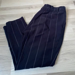 Svarta randiga kostymbyxor - Snygga svarta kostymbyxor med diskreta vita ränder. Byxorna har en klassisk design med hög midja och bälteshällor. Perfekta för en stilren look.