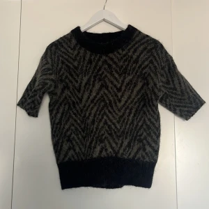 Mönstrad stickad tröja - Säljer en stickad tröja med ett zebra liknande mönster i svart och grön. Tröjan har korta ärmar och en rund halsringning. Perfekt för kyligare dagar!