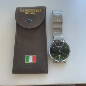 Klocka från Lorenzo Milano med silverarmband - Elegant klocka från Lorenzo Milano med en stilren svart urtavla och italiensk flagga. Klockan har ett silverfärgat mesh-armband och levereras i ett brunt fodral med tryckknapp och italiensk flagga. Ny pris 800kr mitt pris 500kr