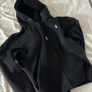 Svart hoodie från Ralph Lauren - Säljer en stilren svart hoodie från Ralph Lauren med dragkedja och huva. Den har en broderad logga på bröstet och är perfekt för en casual look. Passar bra till både jeans och joggers.