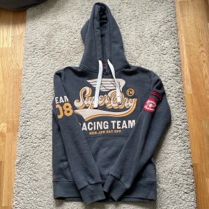 Mörkblå hoodie från Superdry - Säljer en snygg mörkblå hoodie från Superdry med tryck på framsidan och ärmen. Den har en justerbar huva med vita snören och ribbade muddar vid ärmslut och nederkant. Perfekt för en avslappnad stil! Modellen säljs inte längre!!Priset är inte hugget i sten! Träffas helst ( Stockholm )