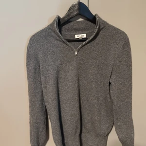 Grå tröja med dragkedja från Soft Goat - Säljer en stilren grå tröja från Soft Goat med dragkedja vid halsen. Tröjan är långärmad och perfekt för kyligare dagar. Den har en klassisk design som passar till det mesta. Perfekt för dig som gillar enkel och tidlös stil.