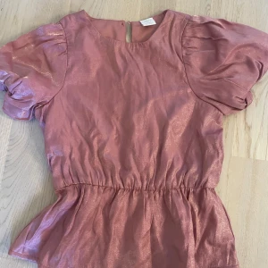 Rosa glittrig topp med puffärmar från Lindex - Säljer en rosa glittrig topp från Lindex med korta puffärmar och resår i midjan. För barn i storlek 128 7/8 år