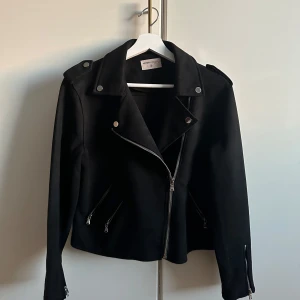 Svart bikerjacka från Ketton Casual - Säljer en svart bikerjacka från Ketton Casual med silvriga dragkedjor och knappar. Jackan har klassisk krage, långa ärmar och flera fickor framtill. Perfekt för en cool och stilren look.