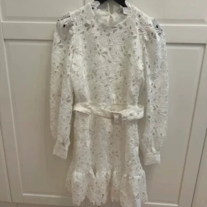 Vit spetsklänning med bälte - Superfin vit klänning i spets med lång ärm och bälte i midjan. Klänningen har blommigt mönster, volangdetaljer nertill och hög hals. Perfekt för somriga tillfällen eller studentfirande.Superfin och true to size! Köpt på vinted men oanvänd av mig och tidigare säljare. Nypris 1100! Pris kan diskuteras vid snabb affär!