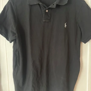 Svart pikétröja från Polo Ralph Lauren - Klassisk svart pikétröja från Polo Ralph Lauren med korta ärmar och vit broderad logga på bröstet. Tröjan har krage och knappar framtill. Perfekt för en stilren och avslappnad look.