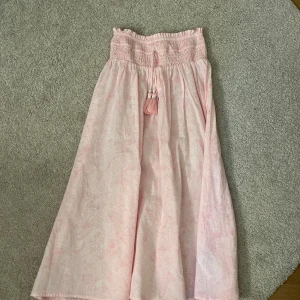 Ljusrosa långkjol med smock och knytband - Supersöt ljusrosa långkjol med smock i midjan och dekorativa knytband med tofsar. Kjolen har ett diskret mönster och är luftig med lätt vidd. Perfekt för varma dagar och enkel att styla med en enkel topp. Säljer eftersom att den blivit för kort. Pris kan diskuteras!💓🫶🏻 Skriv till mig vid frågor.