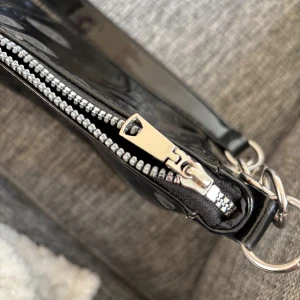 Svart axelväska med kedjedetaljer. Material:lack.   - Snygg svart axelväska i glansigt material med silverfärgad dragkedja och kedjedetaljer på axelremmen. Perfekt accessoar för att lyfta din outfit.