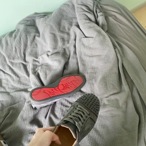 Gråa sneakers med nitar från Christian Louboutin - Säljer ett par gråa sneakers från Christian Louboutin med ikonisk röd sula och nitar framtill. Skorna har snörning och är tillverkade i mocka. Perfekta för dig som vill sticka ut med en lyxig och edgy stil. Box får du med och en nit är av beroende på priset