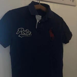 Mörkblå pikétröja från Ralph Lauren - Säljer en vintage mörkblå pikétröja från Polo Ralph Lauren med vit och röd logga på bröstet. Tröjan har klassisk krage med knappslå och korta ärmar. Passar S eller tajt M