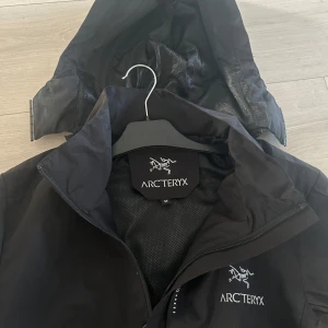 Jacka Arc'teryx svart - Säljer en svart skaljacka från Arc'teryx med huva och dragkedja framtill. Jackan har två sidofickor med dragkedja och justerbar huva. Perfekt för utomhusaktiviteter och skyddar mot vind och regn. Nytt skick