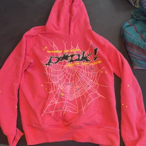 Rosa hoodie med spindelnätsmönster från Spider - Cool rosa hoodie från Spider med stort vitt spindelnätsmönster och text på bröstet. Hoodien har färgglada stjärnor och prickar på ärmarna och framsidan. Tillverkad i bomull och har en klassisk huva och magficka. Pris ej hugget i sten! 