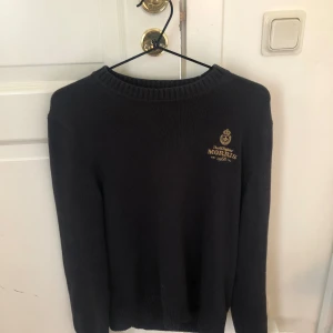 Svart stickad tröja från Morris - En svart stickad tröja från Morris med en broderad logotyp i guld på bröstet. Tröjan har långa ärmar och en ribbad krage, ärmslut och nederkant. Är i storlek XS men passar S. Lite sliter men helt ok skick. Passar mig som är 174 perfekt men passar även 170-175. hör av er vid frågor 