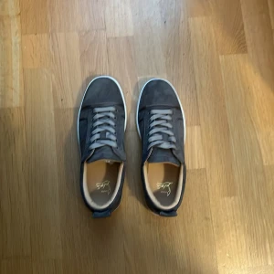 Gråa sneakers i mocka med vit sula - Bara serösa köpare tack!!!   Säljer ett par stilrena gråa sneakers från Louboutin i mjuk mocka med klassisk vit sula och röda undersidor. Skorna har rund tå, snörning och diskreta metallhål på sidorna. Perfekta för dig som gillar en grish/laid back stil.