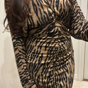 Zebramönstrad omlottklänning - Säljer en snygg zebramönstrad klänning i beige och svart med omlott-effekt och rynkade detaljer längs sidorna. Klänningen har långa ärmar och figurnära passform, perfekt för dig som vill sticka ut.