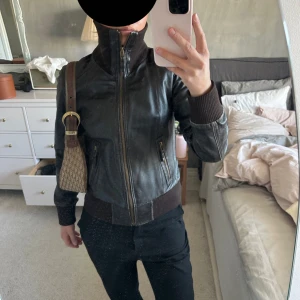 Svart skinnjacka med hög krage - Säljer en svart skinnjacka med hög krage och ribbade muddar i midja och ärmslut. Jackan har dragkedja framtill och två fickor med dragkedja. Snygg och tidlös modell som passar till mycket.