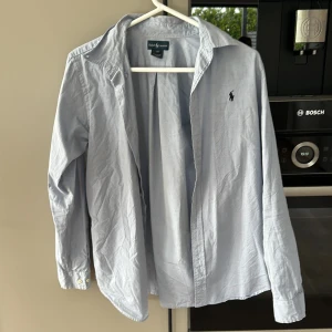 Ljusblå skjorta från Ralph Lauren - Klassisk ljusblå skjorta från Ralph Lauren med broderad logga på bröstet. Skjortan har lång ärm, knappar framtill och är tillverkad i bomull. Perfekt för en stilren och tidlös look.