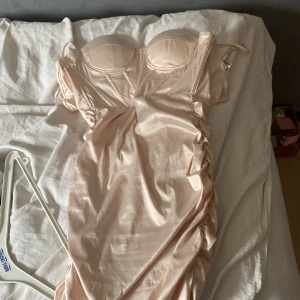 Champagnefärgad satin klänning med kupor - Säljer en snygg axelbandslös klänning i champagnefärgad satin med markerade kupor och rynkade detaljer längs sidorna. Klänningen har en figurnära passform och dragkedja i sidan. Perfekt för dig som vill sticka ut lite extra.