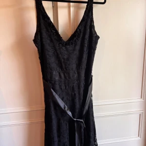 Svart spetsklänning från Oneness - Elegant svart klänning i spets från Oneness med v-ringning och knytband i midjan. Perfekt för en stilfull kväll. Klänningen har en vacker spetsdesign som ger en lyxig känsla.