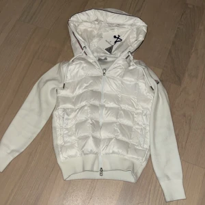 Vit dunjacka från Moncler - Säljer en snygg vit dunjacka från Moncler med huva och dragkedja. Jackan har långa ärmar och en quiltad design framtill. Perfekt för kyligare dagar! Liten fläck från svart ull !denna vara är en replika