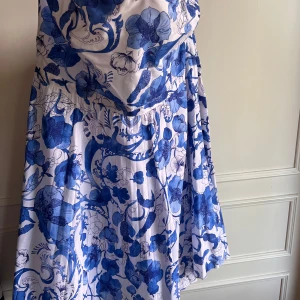 Blåvit blommig klänning från H&M - Säljer en vacker blåvit klänning från H&M med ett blommigt mönster. Klänningen har en elegant passform och är perfekt för sommarens tillställningar. Den har en dragkedja i sidan för enkel påklädning.