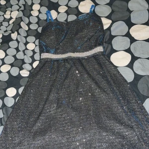 Glittrig svart klänning med blå axelband - Snygg glittrig svart klänning med blå axelband och ett glittrande bälte i midjan. Perfekt för en festlig kväll! Klänningen har en elegant passform och ärmlös design.