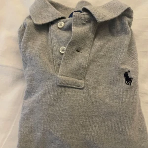 Ralphlauren piké grå - Säljer en klassisk grå pikétröja från Ralph Lauren med korta ärmar och knappar framtill. Tröjan har en broderad logotyp på bröstet och är perfekt för en avslappnad stil. | kostar 2000 retail | passar de som är 150-155. Men även större om man vill att den ska sitta tajt| men också mindre om man vill att den ska sitta normalt.