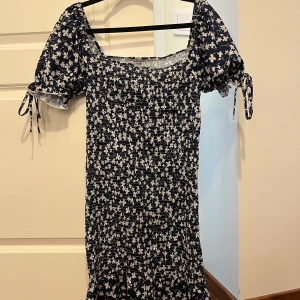 Blommig klänning med puffärmar - Charmig blommig klänning i mörkblått med vita blommor. Den har korta puffärmar med knytband och en smockad design som ger en fin passform. Den kan passa M.