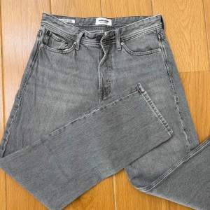 Grå jeans från Jack & Jones - Säljer ett par grå jeans från Jack & Jones med klassisk femficksdesign och raka ben. Jeansen har normal passform och stängs med dragkedja och knapp. Perfekta för en avslappnad stil.