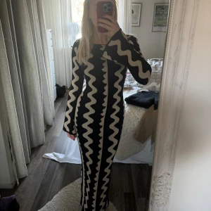 Svart och beige långklänning från NA-KD - Säljer en långärmad stickad långklänning från NA-KD med vågigt mönster i svart och beige. Klänningen har figurnära passform och rund halsringning. Perfekt för dig som vill sticka ut med en unik design.