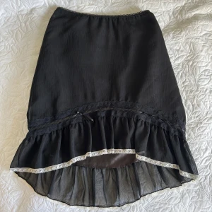 Svart kjol med spetsdetaljer - Snygg svart kjol med spetsdetaljer och volangkant. Perfekt för en stilren look. Kjolen har en elegant design med en liten rosett och transparenta partier vid fållen. Storlek L men insydd för att passa S. 