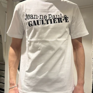 Vit t-shirt från Jean Paul Gaultier - Säljer en vit t-shirt från Jean Paul Gaultier med svart tryck på bröstet. T-shirten har klassisk passform och rund halsringning. Perfekt för dig som gillar stilrena och ikoniska plagg med designer-touch. Nytt pris kostar 2000kr 