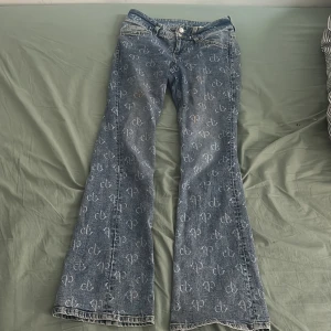 Blå jeans med mönster från H&M - Snygga blå jeans från H&M med ett unikt mönster över hela byxan. De har en bootcut-stil och är perfekta för att ge en cool touch till din outfit. Byxorna har klassisk femficksdesign och knappgylf.