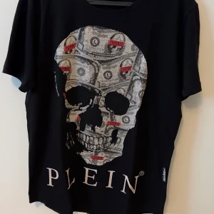 Svart t-shirt med döskalletryck från philipp Plein - Cool svart t-shirt från philipp Plein med stort tryck av en döskalle och sedlar på framsidan. T-shirten har rund halsringning och normal passform. Perfekt för dig som gillar statement-plagg.