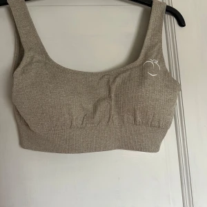 Beige ribbad träningstopp - Beige ribbad träningstopp med breda axelband från märket Peach. Toppen är stretchig och har en croppad passform. 