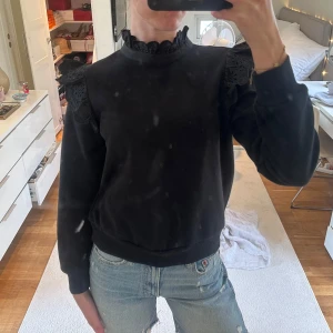 Svart sweatshirt med spetsvolang - Svart sweatshirt med dekorativa spetsvolanger på axlarna och volangkant vid halsen. Tröjan har lång ärm och ribbade muddar vid ärmslut och nederkant. Perfekt för dig som vill ha en basic tröja med extra detaljer.