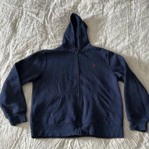 Mörkblå hoodie från Polo Ralph Lauren - Säljer en mörkblå hoodie från Polo Ralph Lauren i storlek XL barn. Men passar som en S. Tröjan har dragkedja, huva och två fickor framtill. Klassisk liten röd logga broderad på bröstet. Perfekt för en avslappnad och stilren look.